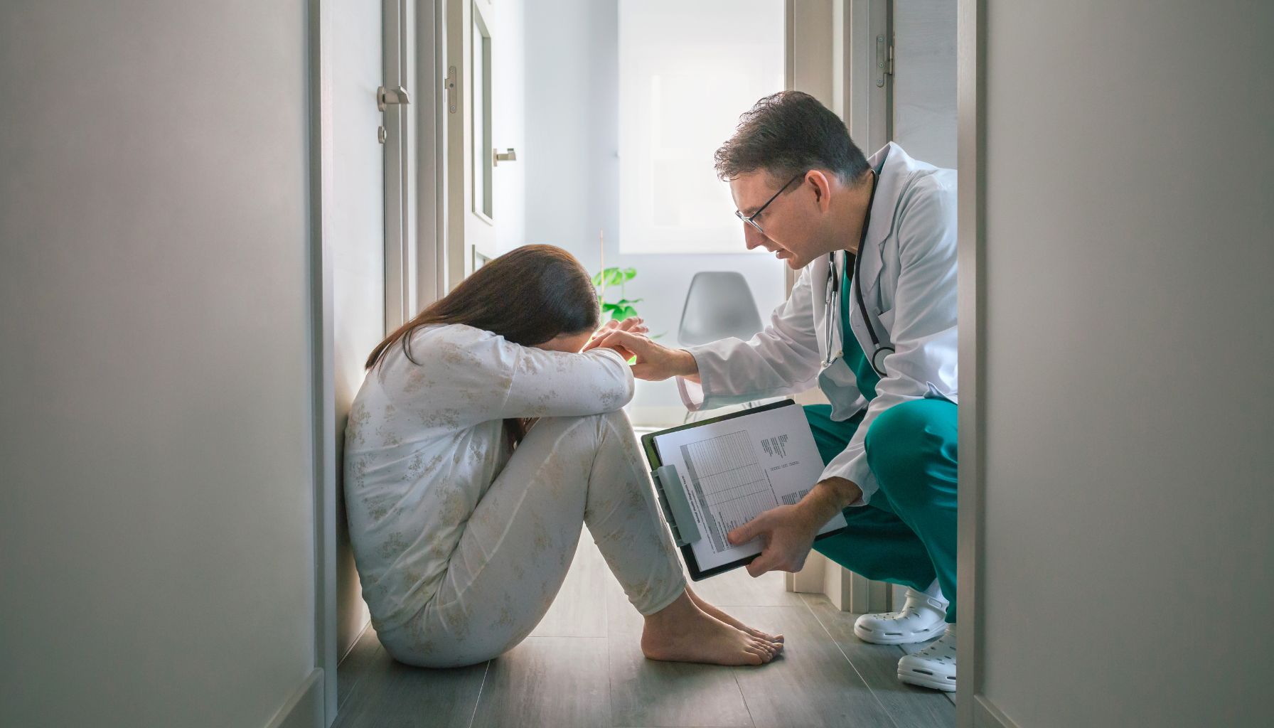 A doctor comforts a seated person in a hallway. The person has their head down and leans against a wall, visibly distressed, possibly worried about water damage restoration needed for their home. The doctor holds a clipboard and places a hand gently on their shoulder. -PureOneServices