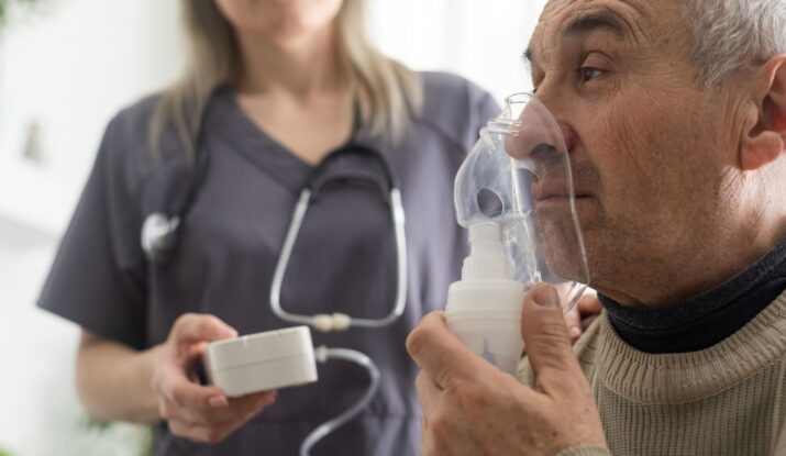 An elderly man uses a nebulizer for respiratory treatment while a healthcare professional stands beside him holding medical equipment, ensuring his well-being amidst ongoing water damage restoration in the facility. -PureOneServices