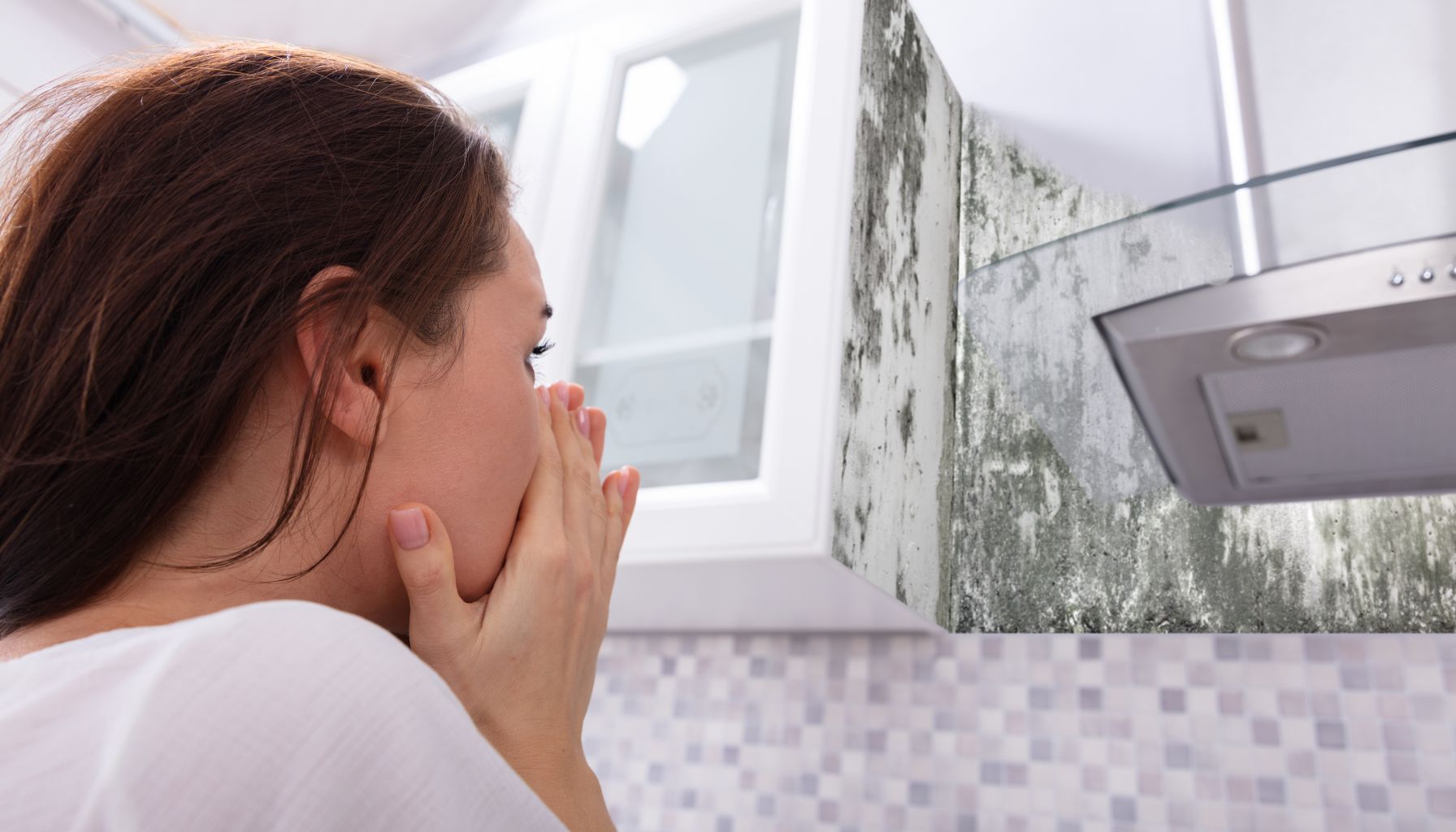 A woman covers her mouth in shock while looking at a mold-covered corner above a kitchen countertop, recognizing the urgent need for mold restoration. -PureOneServices