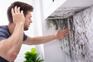 A man looks at a large patch of mold on a kitchen wall, touching his head in apparent frustration or concern. The overwhelming need for water damage restoration is evident, making the situation even more disheartening. -PureOneServices