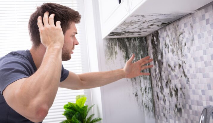 A man looks at a large patch of mold on a kitchen wall, touching his head in apparent frustration or concern. The overwhelming need for water damage restoration is evident, making the situation even more disheartening. -PureOneServices