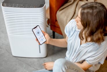 A person sits on the floor near a dehumidifier, holding a smartphone displaying a control app for the device, ensuring effective mold restoration. -PureOneServices