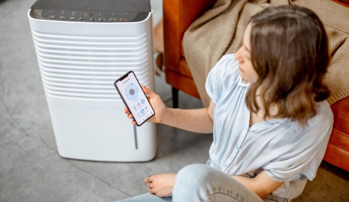 A person sits on the floor near a dehumidifier, holding a smartphone displaying a control app for the device, ensuring effective mold restoration. -PureOneServices