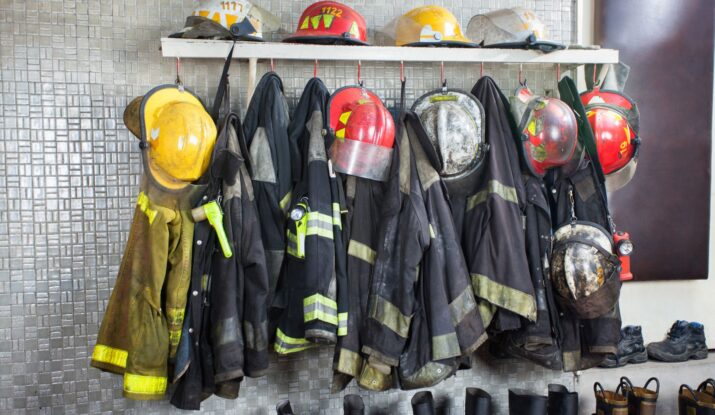 Firefighter helmets and jackets hang on hooks above a row of boots, arranged neatly against a tiled wall, ready for swift action, even in water damage restoration scenarios. -PureOneServices