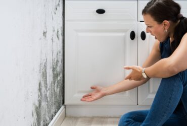 A woman in casual clothes sits on the floor, pointing distressedly at a wall with significant mold growth next to a white cabinet, in desperate need of mold restoration. -PureOneServices