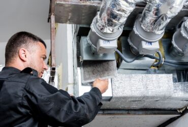 Person in a black uniform inspecting ductwork with multiple pipes and insulation in an HVAC system, likely assessing the need for water damage restoration. -PureOneServices