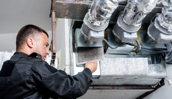 Person in a black uniform inspecting ductwork with multiple pipes and insulation in an HVAC system, likely assessing the need for water damage restoration. -PureOneServices