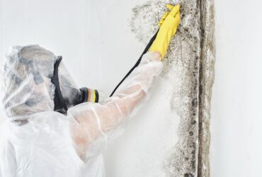 A person in protective gear and a respirator is performing mold restoration by carefully cleaning mold from a wall with a sponge. -PureOneServices