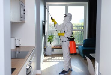 A person in a white protective suit, yellow gloves, and face mask sprays pesticide inside a modern apartment, possibly addressing mold restoration. They are holding a red and black sprayer with a hose pointed at the wall. -PureOneServices