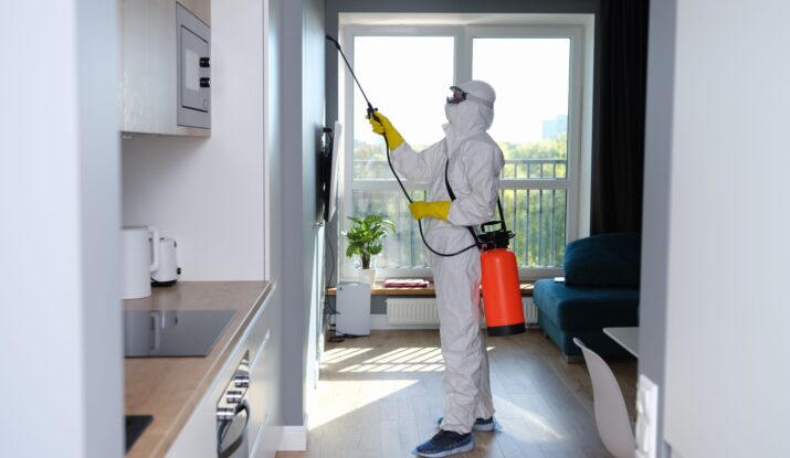 A person in a white protective suit, yellow gloves, and face mask sprays pesticide inside a modern apartment, possibly addressing mold restoration. They are holding a red and black sprayer with a hose pointed at the wall. -PureOneServices