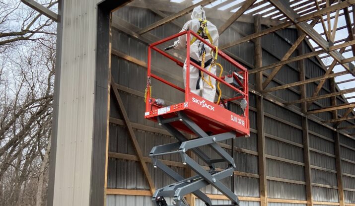 A worker in protective gear stands on a raised scissor lift, inspecting or working on the structure of a large, partially open building, possibly focusing on mold restoration. -PureOneServices