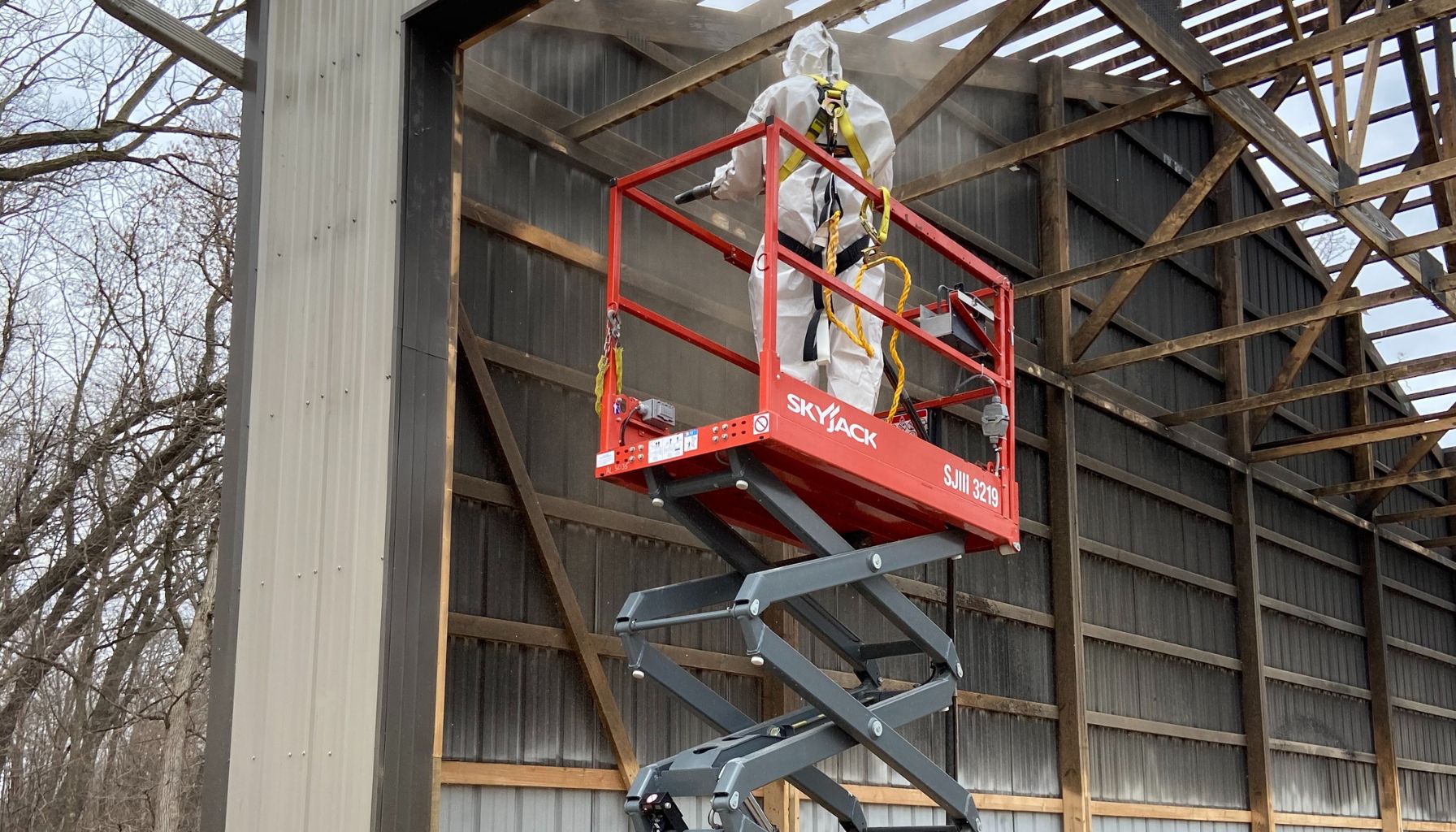 A worker in protective gear stands on a raised scissor lift, inspecting or working on the structure of a large, partially open building, possibly focusing on mold restoration. -PureOneServices