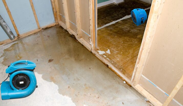 A room with partially constructed walls and a wet concrete floor shows clear signs of water damage restoration. Two blue fans are placed on the ground to help dry the area. A door leads to another room undergoing a similar restoration process. -PureOneServices