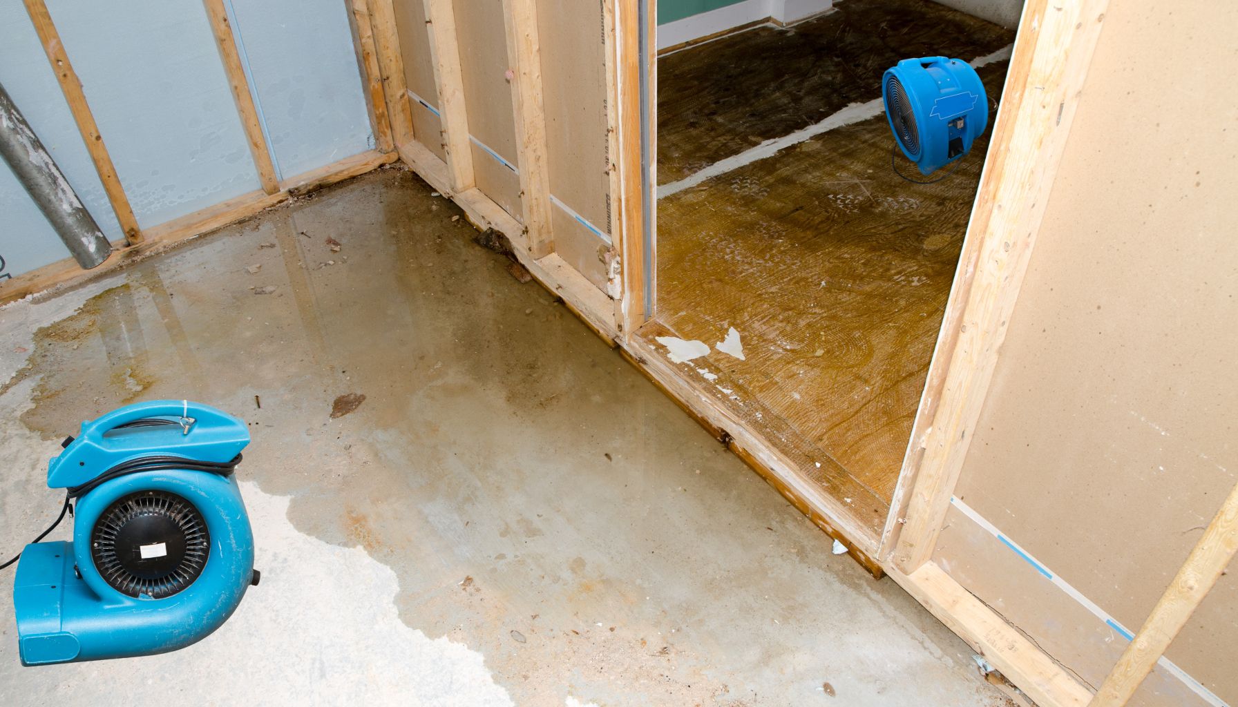 A room with partially constructed walls and a wet concrete floor shows clear signs of water damage restoration. Two blue fans are placed on the ground to help dry the area. A door leads to another room undergoing a similar restoration process. -PureOneServices
