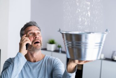 A man on the phone holds a metal bucket to catch water leaking from the ceiling in a modern living room, clearly in need of immediate water damage restoration. -PureOneServices