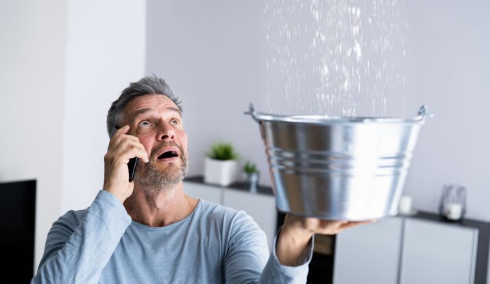 A man on the phone holds a metal bucket to catch water leaking from the ceiling in a modern living room, clearly in need of immediate water damage restoration. -PureOneServices