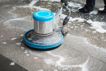 A floor cleaning machine is being used to clean a tile floor, creating soapy water and foam. Nearby, a person in black pants and black shoes stands by, ready for mold restoration after addressing potential water damage restoration needs. -PureOneServices
