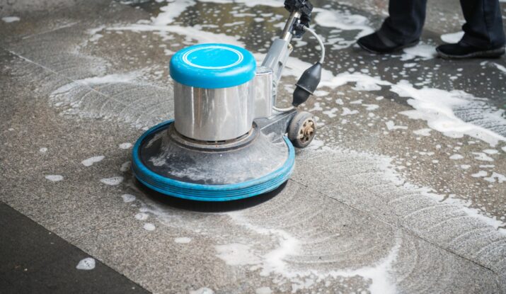 A floor cleaning machine is being used to clean a tile floor, creating soapy water and foam. Nearby, a person in black pants and black shoes stands by, ready for mold restoration after addressing potential water damage restoration needs. -PureOneServices