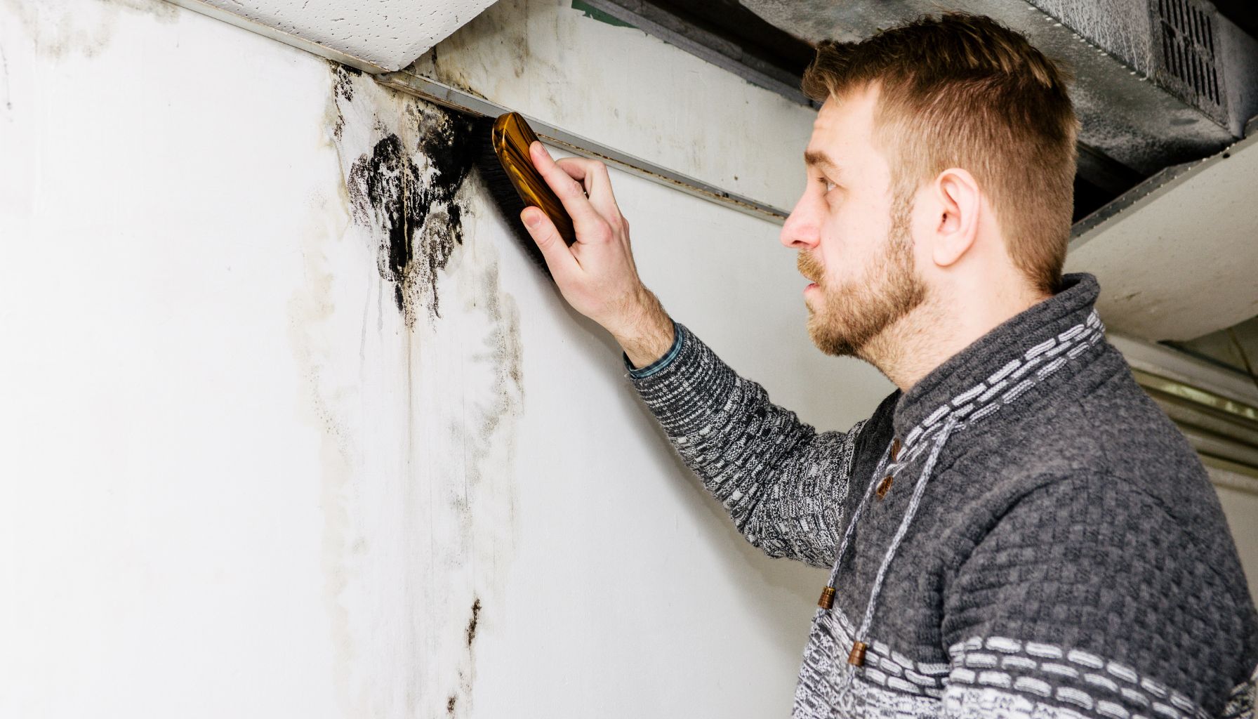 A man in a gray sweater is holding a moisture meter against a white wall with visible water damage and mold, assessing the extent for mold restoration. -PureOneServices
