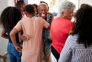 A group of people, including older adults and children, are gathered indoors and hugging each other. One man in the center is smiling broadly, relieved after a successful mold restoration brought their home back to life. -PureOneServices