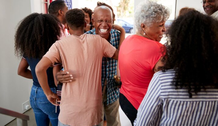 A group of people, including older adults and children, are gathered indoors and hugging each other. One man in the center is smiling broadly, relieved after a successful mold restoration brought their home back to life. -PureOneServices