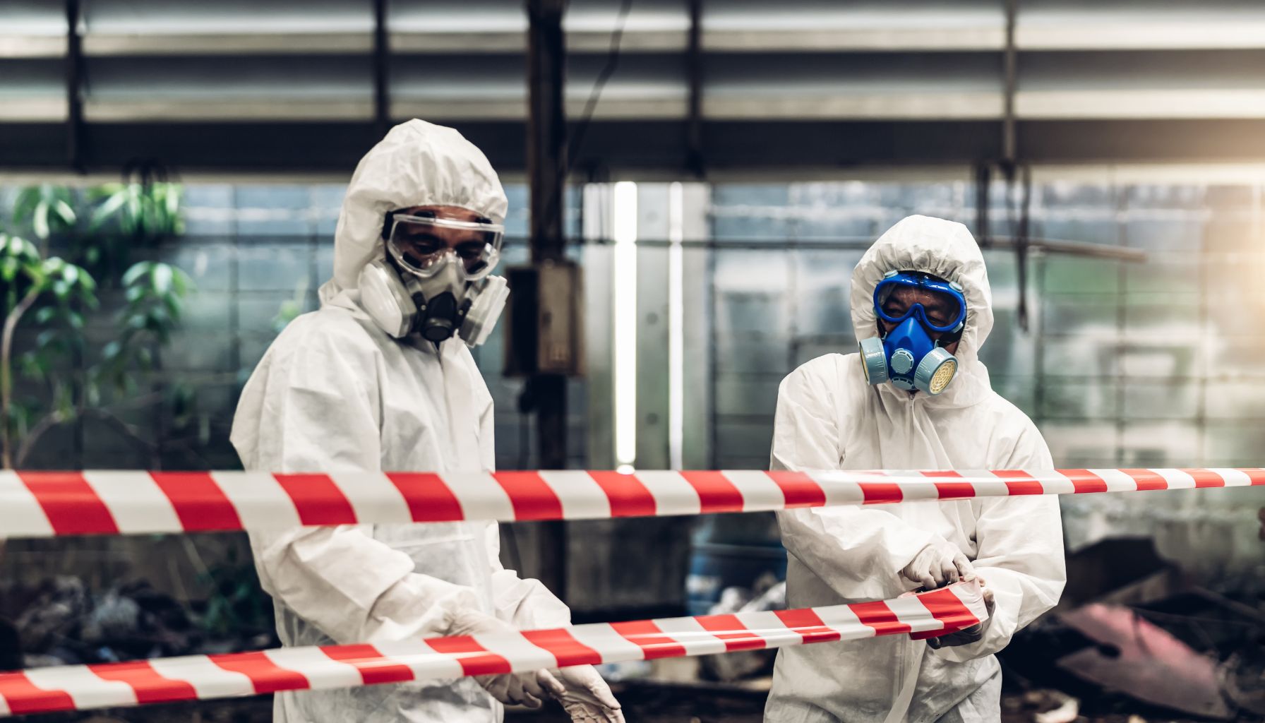 Two people in protective suits and gas masks handle red and white caution tape in an industrial setting, likely engaged in mold restoration efforts. -PureOneServices