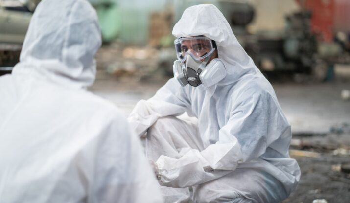 Two individuals in full hazmat suits and respiratory masks are engaged in conversation in an industrial setting, likely discussing water damage restoration. -PureOneServices