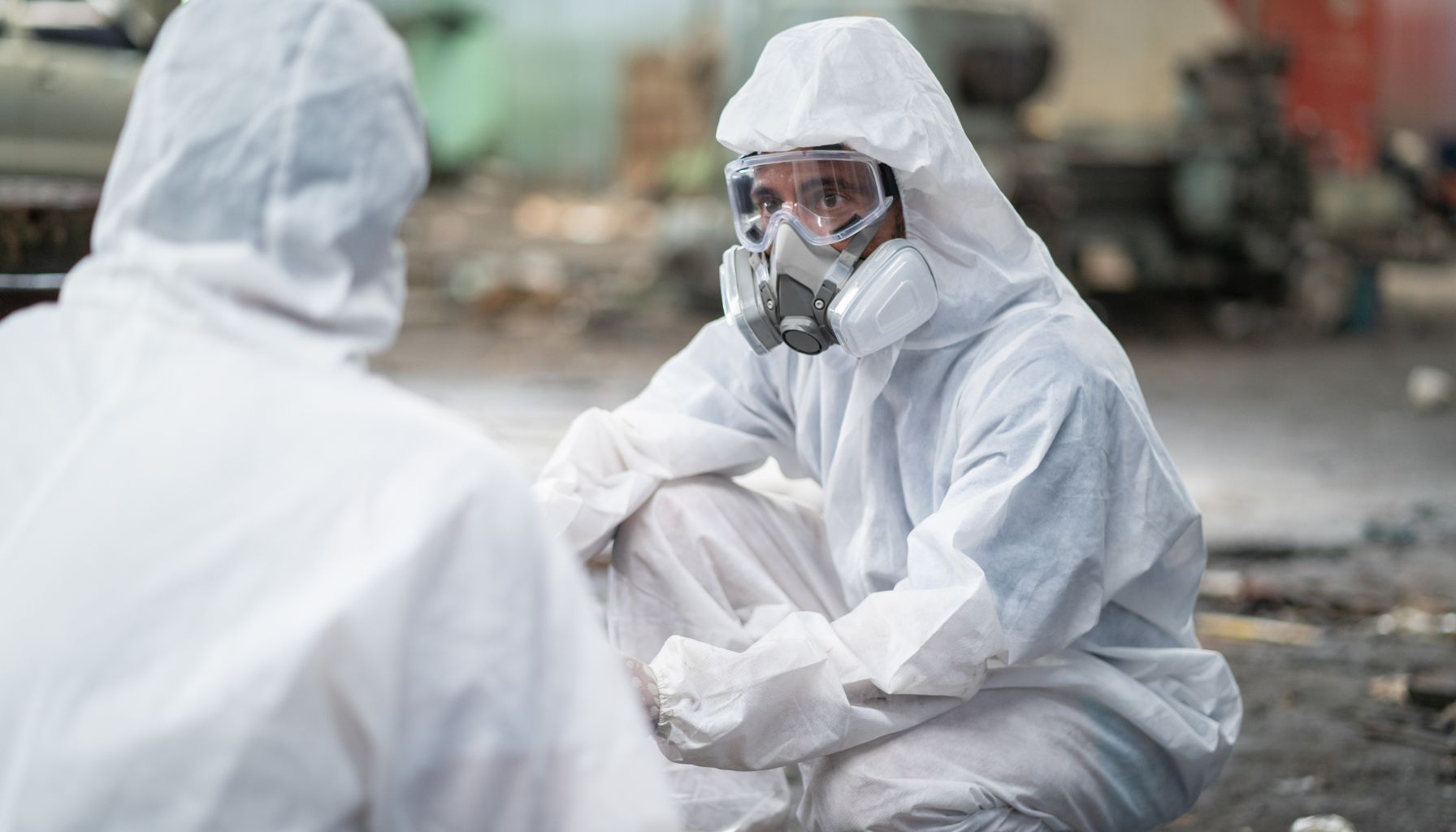 Two individuals in full hazmat suits and respiratory masks are engaged in conversation in an industrial setting, likely discussing water damage restoration. -PureOneServices