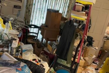 A cluttered room with various items, including books, clothes, boxes, and papers scattered on the floor and surfaces. Shelves and a table are also filled with miscellaneous objects. Curtains are partially drawn, hinting at the need for water damage restoration to address any potential mold issues in the space. -PureOneServices