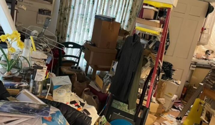 A cluttered room with various items, including books, clothes, boxes, and papers scattered on the floor and surfaces. Shelves and a table are also filled with miscellaneous objects. Curtains are partially drawn, hinting at the need for water damage restoration to address any potential mold issues in the space. -PureOneServices