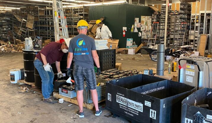 Workers in a warehouse are sorting and handling various items, including batteries and metal barrels. Two of them are wearing hard hats. The warehouse shelves, filled with materials and boxes, include supplies for mold restoration efforts. -PureOneServices