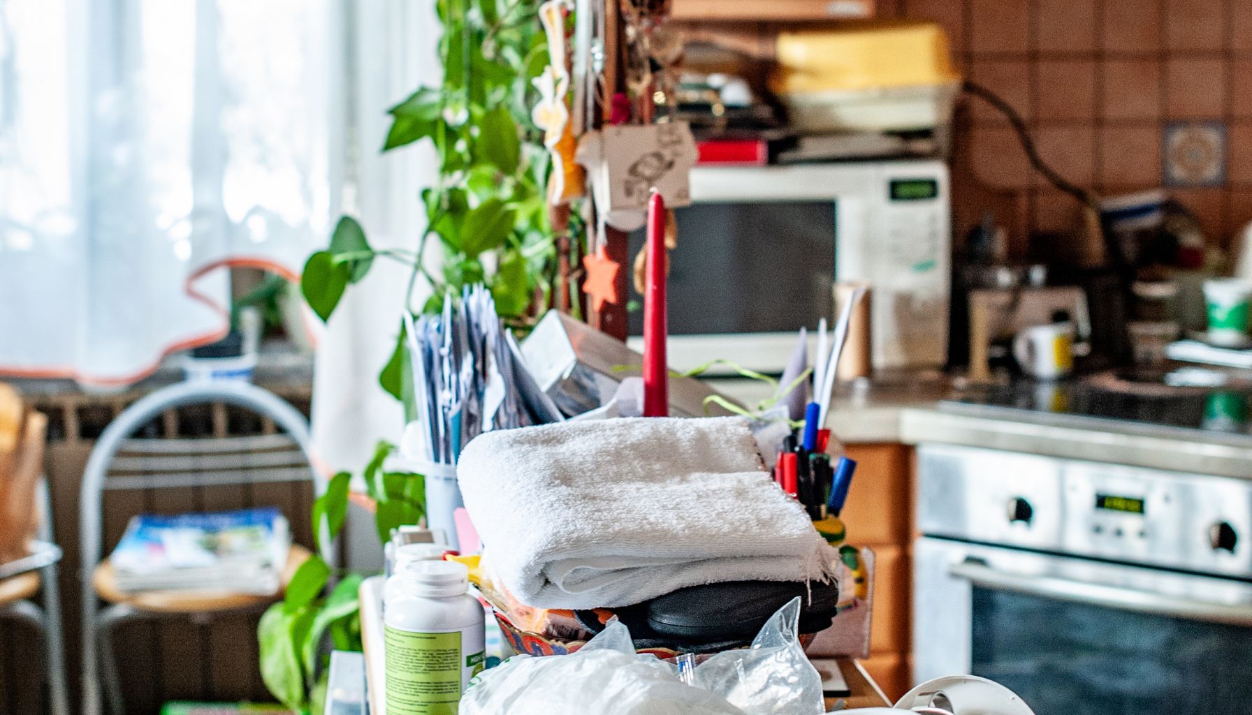 A cluttered kitchen featuring a pile of towels, plants, various kitchen appliances including a microwave and an oven, and other miscellaneous items scattered around the countertop, some of which hint at the need for mold restoration. -PureOneServices