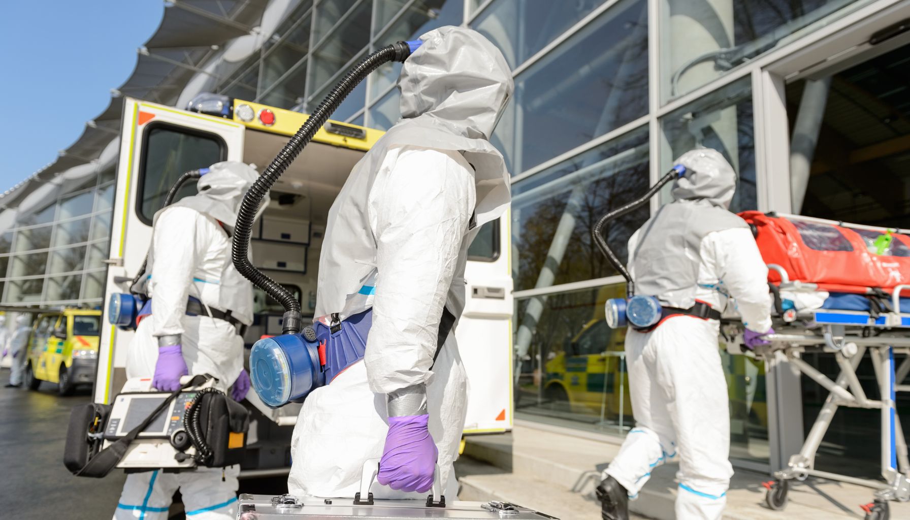 Three individuals in hazmat suits transport equipment and a stretcher into a building from an ambulance parked outside, ready for mold restoration. -PureOneServices