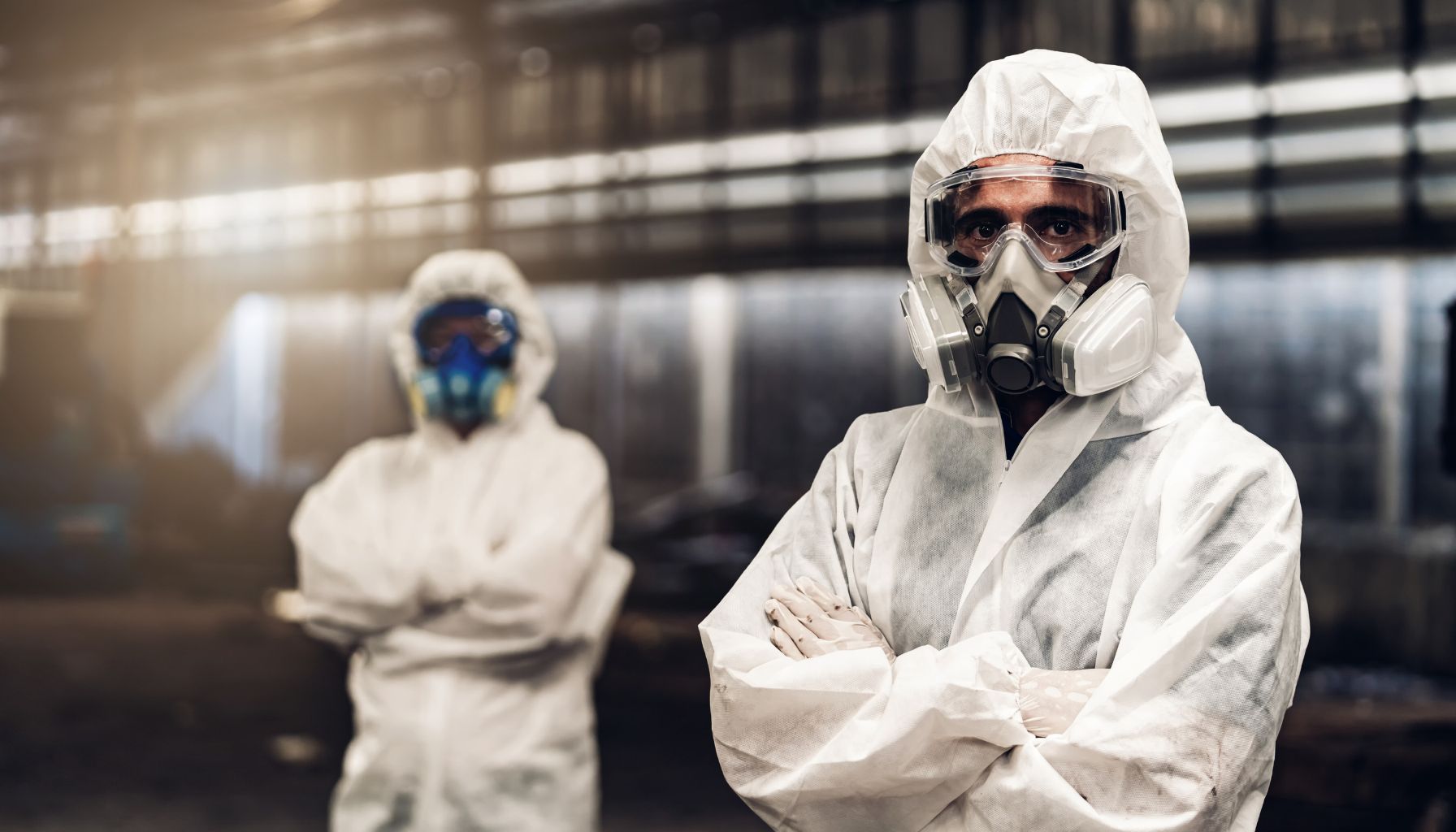 Two individuals wearing white hazmat suits and respirators stand in an industrial setting, one in the foreground with arms crossed, overseeing mold restoration, and another in the background. -PureOneServices