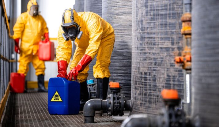 Two workers in yellow hazmat suits and gas masks handle containers with biohazard symbols in an industrial setting, possibly related to mold restoration. One worker pours from a red container, while the other handles a blue container. -PureOneServices