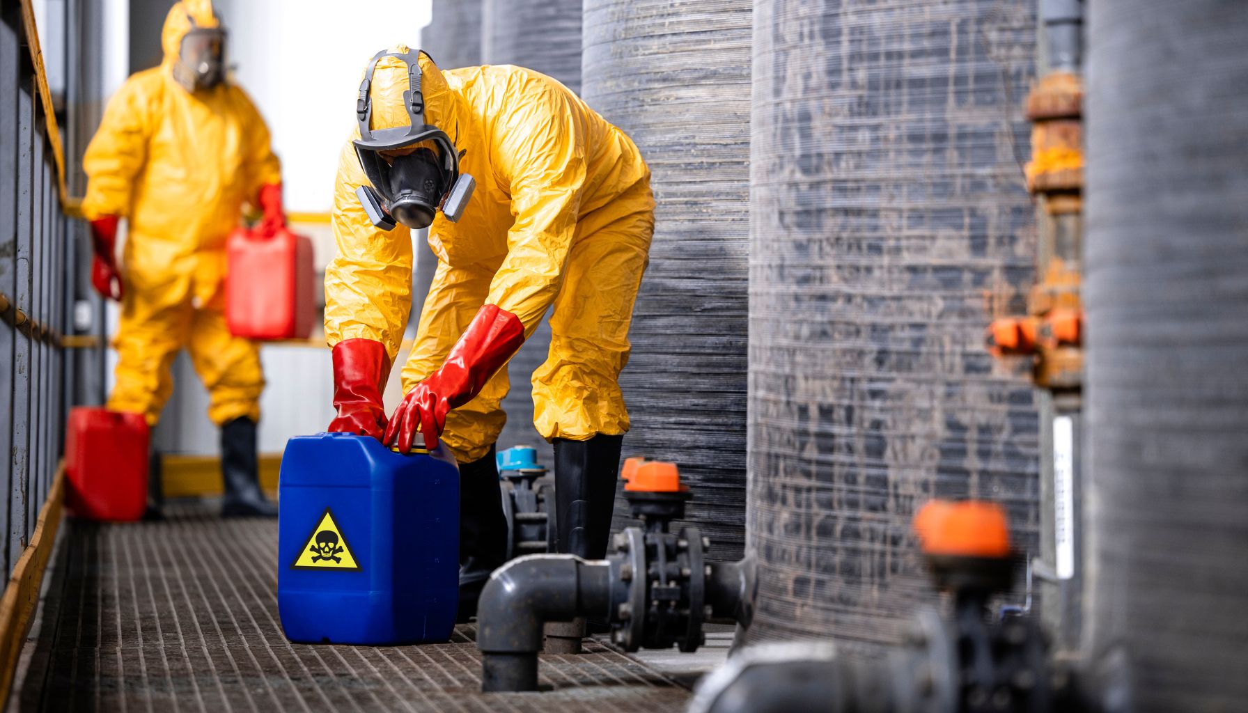 Two workers in yellow hazmat suits and gas masks handle containers with biohazard symbols in an industrial setting, possibly related to mold restoration. One worker pours from a red container, while the other handles a blue container. -PureOneServices
