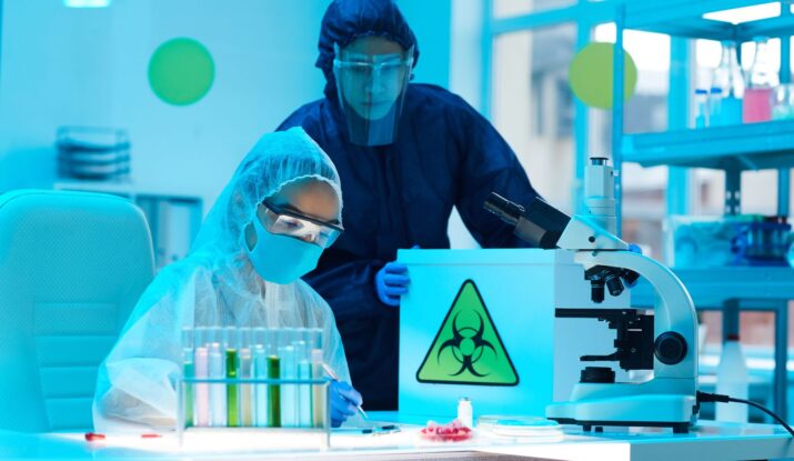 Two scientists in protective suits and masks are working in a laboratory with test tubes and a microscope. One holds a container with a biohazard symbol, crucial for their mold restoration research. -PureOneServices