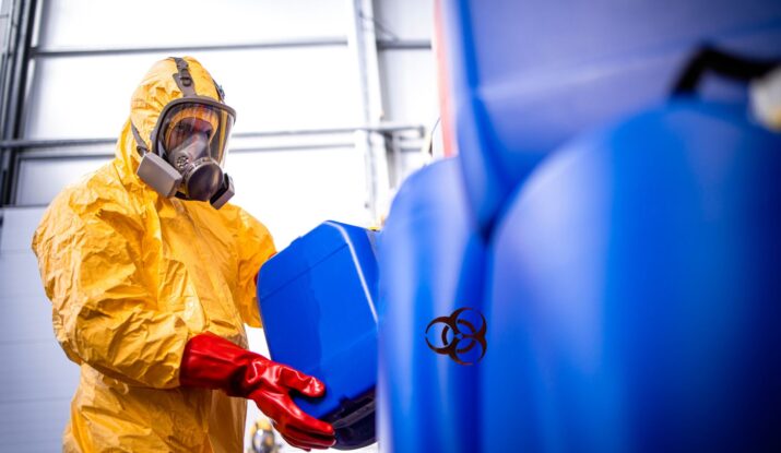 A person in a yellow hazmat suit and red gloves carefully handles a blue container amidst others, marked with a biohazard symbol, during mold restoration. -PureOneServices