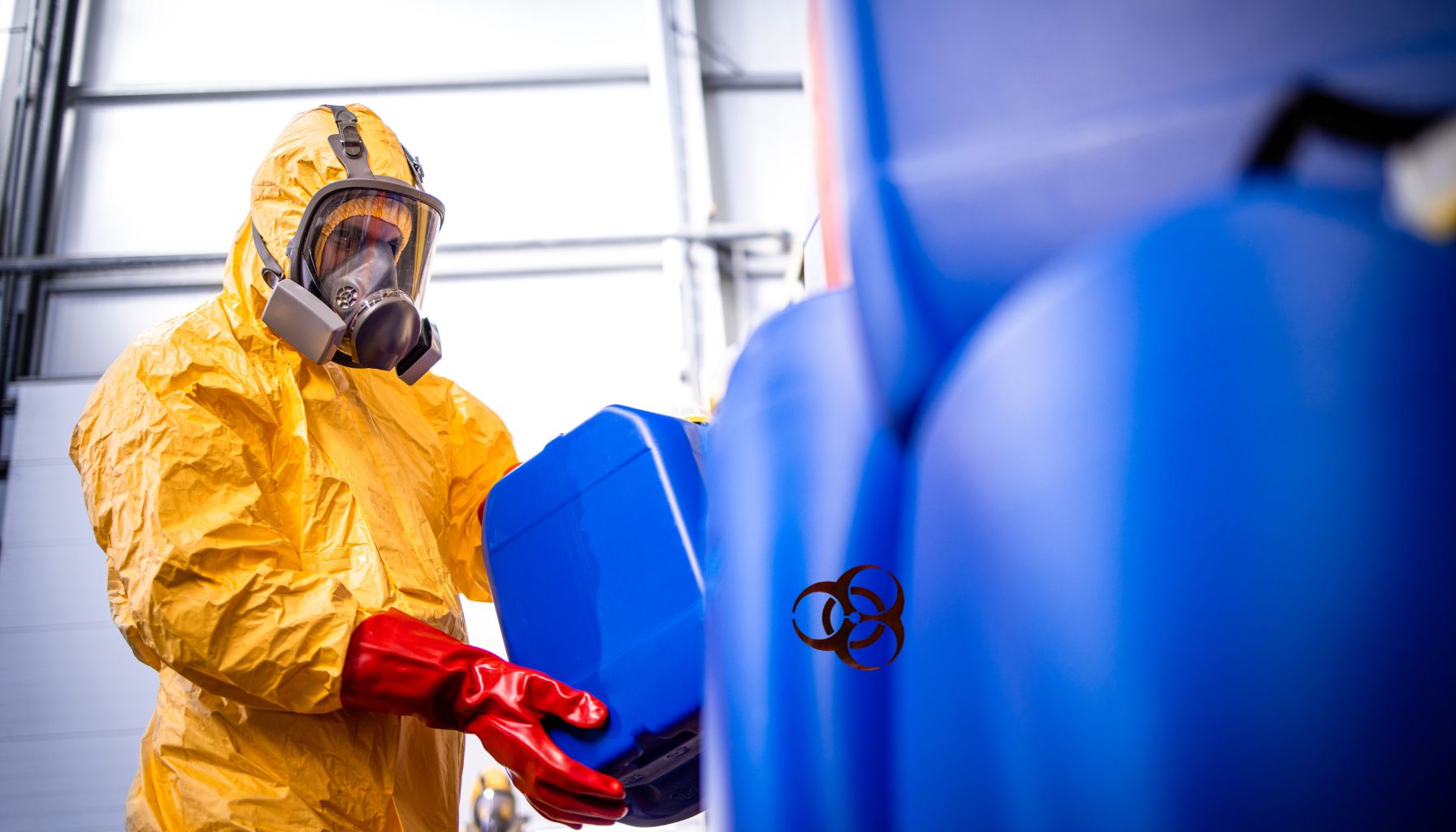 A person in a yellow hazmat suit and red gloves carefully handles a blue container amidst others, marked with a biohazard symbol, during mold restoration. -PureOneServices